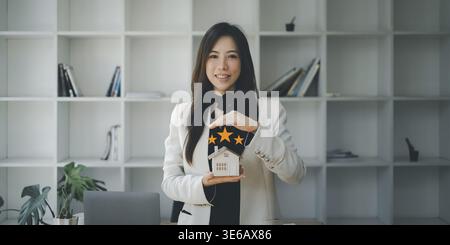 Asiatischer Versicherungsvertreter macht Handsymbol, um das Haus vor einem Unfall zu schützen. Immobilien- und Vertragskonzept Stockfoto
