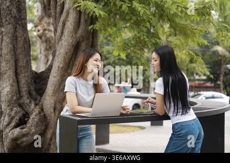 Zwei verschiedene Studentinnen diskutieren Studienmaterial, während sie einen Laptop an einem Tisch im Freien in der Nähe eines Baumes auf dem Universitätsgelände benutzen Stockfoto