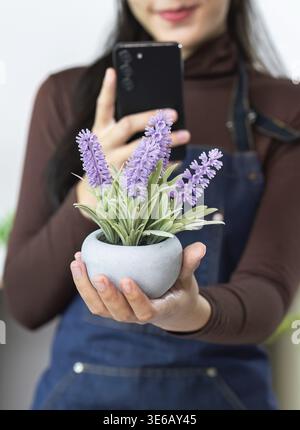 Eine junge Frau hält eine Topfpflanze, während sie mit ihrem Smartphone ein Foto macht, was ihre Leidenschaft für die Gartenarbeit unterstreicht Stockfoto