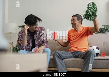 Ein älterer Mann, der fröhlich Pompons und eine rote Tasse hält, um ein Fußballspiel zu feiern, während ein junger Mann nachdenklich aussieht und beide einen Moment in ihrer Lebensführung teilen Stockfoto