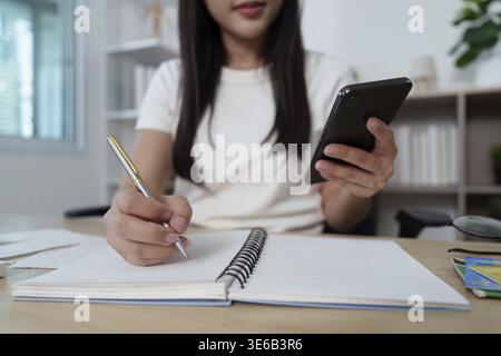 Eine junge Frau schreibt in ihrem Notebook, während sie ein Smartphone hält, und betont dabei die Integration von Technologie in die tägliche Planung Stockfoto
