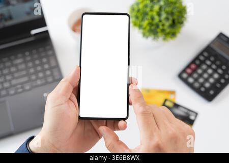 Nahaufnahme der Hände mit leerem Smartphone-Bildschirm über einem Schreibtisch mit Laptop, Rechner, Karten, Kaffee und Pflanze geeignet für App-Design, Stockfoto
