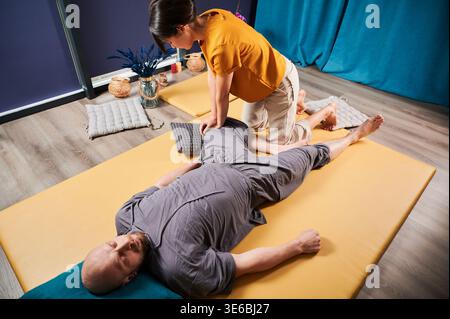 Masseurin, die thai-Yoga-Massage macht. Eine Therapeutin, die traditionelle Massagebehandlung durchführt und den Körper des männlichen Patienten dehnt. Glatzköpfiger Kunde liegt auf der Matte und genießt eine Handmassage in ruhiger Atmosphäre. Stockfoto