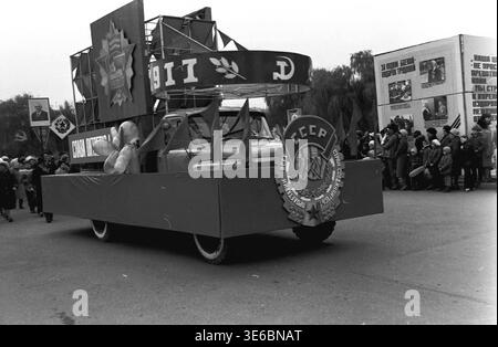 Slawiansk, 7. November 1983, Platz der Oktoberrevolution (heute Soborna). Ein Archivfoto, das einen dekorierten Wagen bei einer Parade zeigt. Der Schwimmer verfügt über einen großen Ring mit „1917“ sowie einen Hammer und eine Sichel. Auf der Vorderseite zeigt ein riesiges sowjetisches Staatswappen den russischen Text: 'PROLETARII VSEKH STRAN, SOEDINYAYTES'!' (Proletarier aller Länder, vereinigt euch!) Zuschauer stehen auf dem Bürgersteig und beobachten die Demonstration am großen Informationswürfel. Das ist eine Szene des friedlichen Lebens im Donbass. Stockfoto