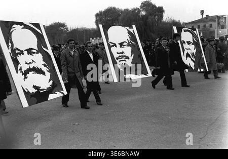 Slawiansk, 7. November 1983, Platz der Oktoberrevolution (heute Soborna). Ein Schwarzweiß-Archiv, das während der Parade eine Demonstrationskolumne aufnimmt. Männer in Mänteln und Anzügen marschieren aktiv und tragen große grafische Porträts kommunistischer Führer Karl Marx, Wladimir Lenin und Friedrich Engels auf Holzpfählen. Weinende Weidenbäume, Stadtgebäude und Massen anderer Märsche sind im Hintergrund deutlich zu sehen. Eine lebendige Szene der sowjetischen Staatsideologie. Das ist eine Szene des friedlichen Lebens im Donbass. Stockfoto