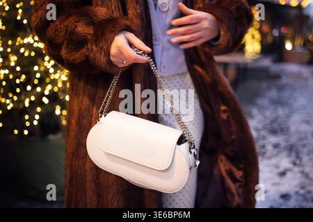 Frau in einem braunen Pelzmantel trägt eine weiße Handtasche im Freien, mit verschwommenen festlichen Lichtern, die ein warmes Ambiente im Winter schaffen Stockfoto