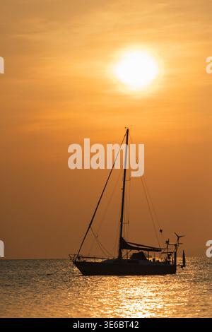 Silhouette einer Segelyacht, die auf ruhigem Meer bei goldenem Sonnenuntergang mit heller Sonne über dem Horizont und glitzernden Wasserreflexionen verankert ist Stockfoto