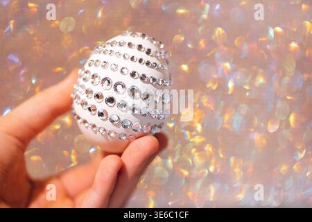 Weißes Osterei mit funkelnden Strasssteinchen wird in der Hand gehalten und vor einem unscharfen, farbenfrohen Hintergrund mit schimmerndem Effekt gesetzt Stockfoto