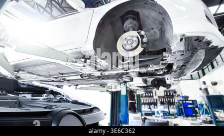 Unterboden eines Fahrzeugs auf einer hydraulischen Hebebühne, Ein niedriger Winkel eines weißen Autos, das auf einer hydraulischen Hebebühne in einer modernen Werkstatt angehoben wurde, mit Bremsscheiben, Fahrgestell, und Stockfoto