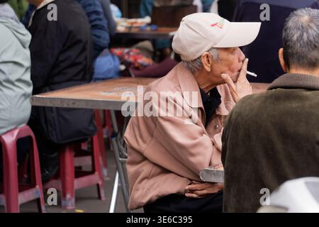 Taipei, Taiwan - 13. März 2024: Ein älterer Taiwaner macht eine Rauchpause am Tisch im Freien nahe dem Cisheng Tempel, zwischen roten Stühlen und verschwommenen Gästen. Stockfoto