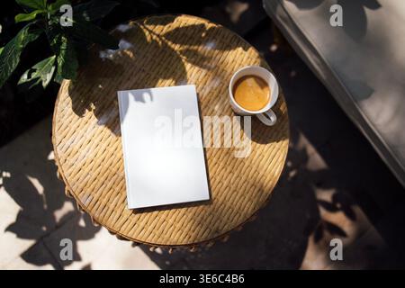 Das leere Notizbuch liegt auf einem runden Korbtisch neben einer weißen Kaffeetasse, die mit Kaffee gefüllt ist, mit grünen Blättern und sanften Schatten, die eine ruhige Outdooratmosphäre schaffen Stockfoto