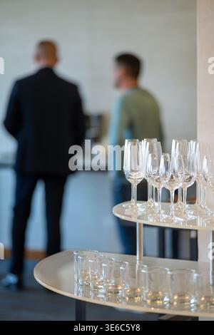 Leere Weingläser und Gläser auf einem zweistöckigen Stand bereit für eine Party oder Veranstaltung, mit verschwommenen Personen im Hintergrund. Stockfoto