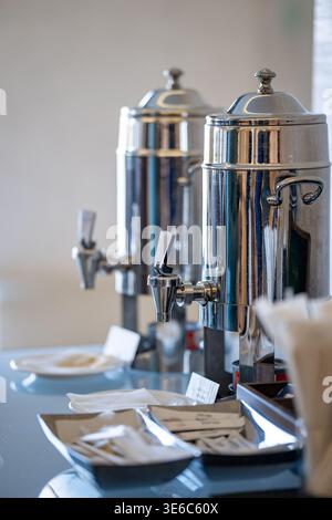 Selbstbedienungs-Kaffeestation mit glänzenden Edelstahl-Getränkeurnen, Zuckerpackungen, Servietten und Keksen auf einem Tisch. Stockfoto