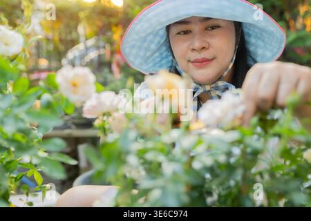 Glückliche asiatische Frau mit blauem Hut, die tagsüber ihre Pflanzen im Garten pflegt. Frau im Garten, selektiver Fokus. Stockfoto