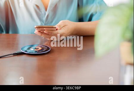 Smartphone auf der Hand der Frau mit einem Ladepad auf Holztisch. Mobiltelefon in der Nähe des kabellosen Ladegeräts. Modernes Lifestyle-Technologiekonzept. Stockfoto