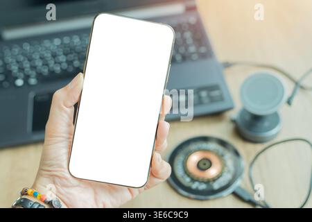 Frauenhand mit Smartphone mit leerem weißen Bildschirm auf verschwommenem Bild eines drahtlosen Ladegeräts am Schreibtisch mit Laptop-Hintergrund. Kopierbereich. Stockfoto