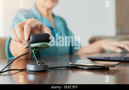 Frau mit Ohrhörern, die auf dem Wireless-Pad neben dem Smartphone auf dem Wireless-Ladegerät am Schreibtisch aufgeladen werden. Selektiver Fokus. Modernes Konzept für Arbeitsplatzgeräte. Stockfoto