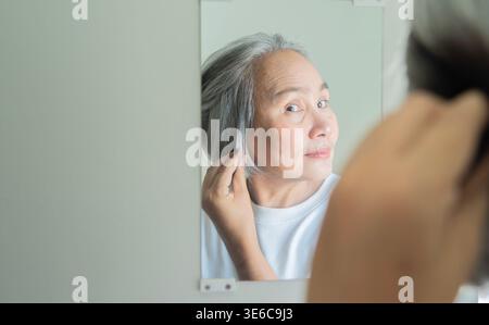 Asiatische schöne und lächelnde Frau mittleren Alters, die ihr graues Haar in der Nähe des Spiegels mit Kopierraum berührt. Selektiver Fokus. Graues Haar Lifestyle Concep Stockfoto