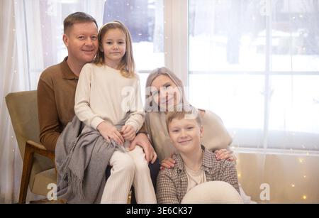 Eine vierköpfige Familie, darunter zwei Kinder, posiert in Winterkleidung drinnen, mit einer verschneiten Szene vor dem Fenster und sanfter Beleuchtung, die den gemütlichen Lärm unterstreicht Stockfoto