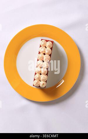Rechteckige Scheibe roter Samtkuchen mit Schlagsahne und roten Streuseln, serviert auf einem gelb-weißen Teller über einer weißen Tischdecke. Stockfoto