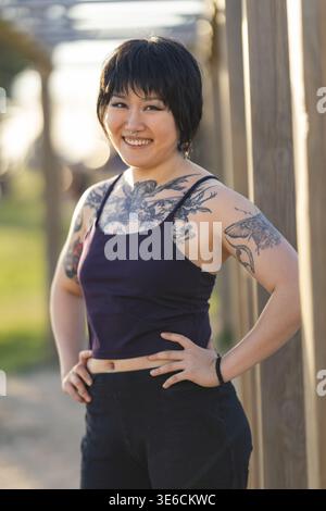 Junge asiatische Frau mit Brust- und Armtattoos lächelnd, während sie mit Händen auf Hüften posiert Stockfoto