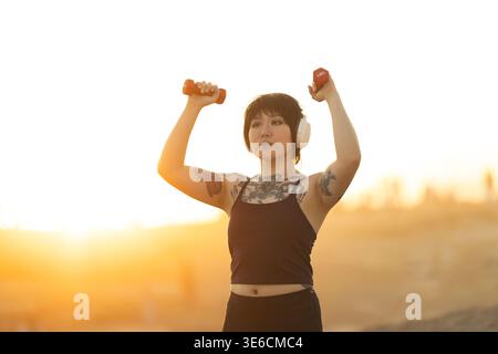 Frau, die während der goldenen Stunde ein Outdoor-Workout mit Kurzhanteln mit Kopfhörern durchführt Stockfoto