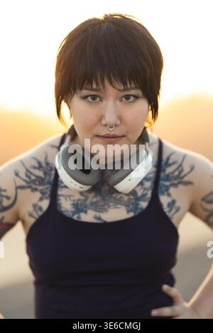 Junge Frau mit Kopfhörern und Brusttattoos, die sich bei Sonnenuntergang selbstbewusst im Freien posiert Stockfoto