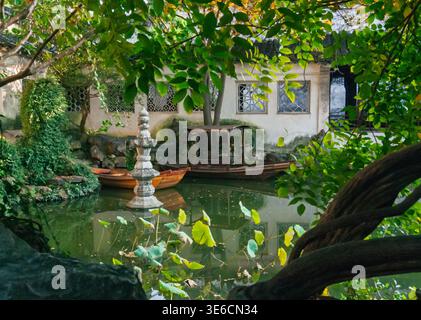 Ruhiger chinesischer Gartenteich mit kleinen Holzbooten und einer dekorativen Steinpagode an einem traditionellen Gebäude an einem klaren Tag Stockfoto