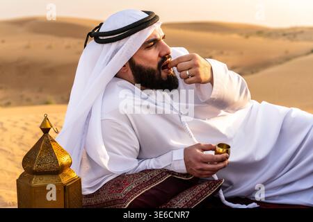Junger Mann aus dem Nahen Osten, der traditionelle Kandura und Gutra trägt und sich auf einem Teppich in der Wüste entspannt, Datteln isst und einen kleinen goldenen Becher in der Nähe einer de hält Stockfoto