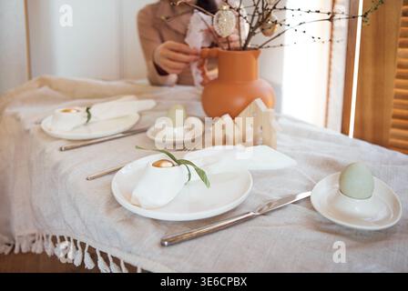 Esstisch mit weißen Tellern, dekorativen Eiern und einer Vase mit Zweigen, die eine gemütliche Atmosphäre in einem Innenbereich mit Holzjalousien schaffen Stockfoto