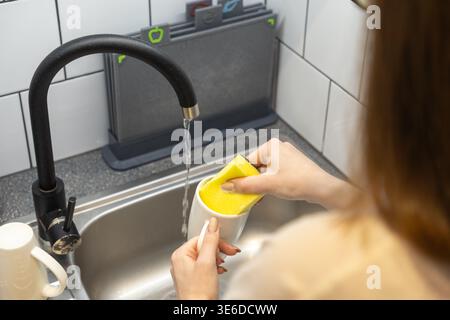 Nahaufnahme von weiblichen Händen, die einen weißen Keramikbecher mit einem leuchtend gelben Scheuerschwamm in einem Edelstahlwaschbecken reinigen, wodurch die praktische h Stockfoto