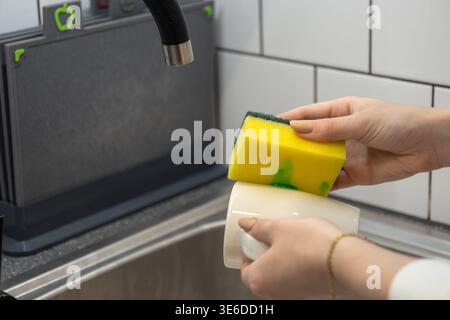 Nahaufnahme von Händen, die eine weiße Tasse mit einem matschigen gelben Schwamm und einem grünen Flüssigwaschmittel sorgfältig schrubben und so ein effizientes Geschirrspüler betonen Stockfoto