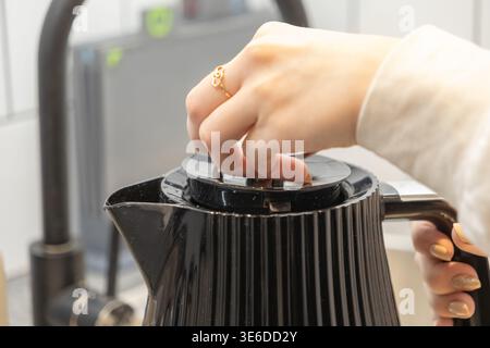 Eine Nahaufnahme der Hand, die den eleganten schwarzen Deckel des Wasserkochers öffnet, um Wasser hinzuzufügen, und die Zubereitung von Tee oder Kaffee in einem modernen Zuhause vorbereitet Stockfoto
