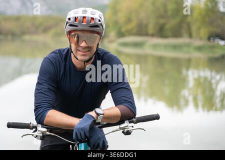 Radfahrer lächeln auf dem Mountainbike an einem ruhigen See mit Helm, Handschuhen und Sportsonnenbrille Stockfoto