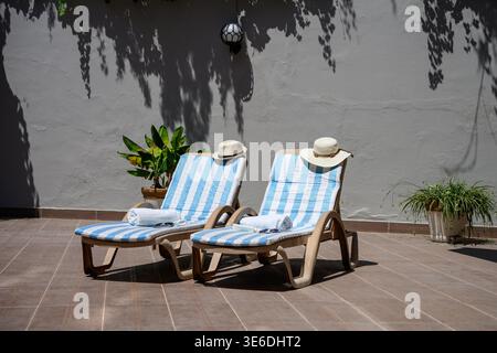 Zwei leere Sonnenliegen mit Strohhüten und Handtüchern auf einer sonnigen Terrasse. Sommerurlaub, Entspannung und Luxus-Resort-Konzept, Reisen. Minimalistisches Outdoo Stockfoto