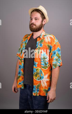 Studio-Porträt eines jungen kaukasischen Mannes mit Bart, der ein leuchtendes orangenes Aloha-Hemd, ein anthrazitfarbenes T-Shirt und einen Strohhut trägt. Ideal für den Sommer Stockfoto