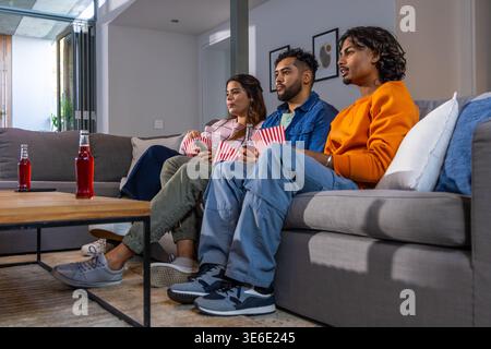 Freunde sitzen zu Hause auf einem grauen Sofa und schauen Filme, während sie Popcorn-Boxen halten Stockfoto