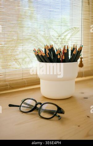 Farbstifte im Halter und Lesebrille auf einem Holztisch am Fenster Stockfoto