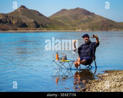 Ein bärtiger Mann trinkt Kaffee im Wasser, während er auf einem Campingstuhl sitzt. Ein glücklicher junger Mann hat an einem warmen Frühlingstag im Wasser ein Lager aufgelegt Stockfoto