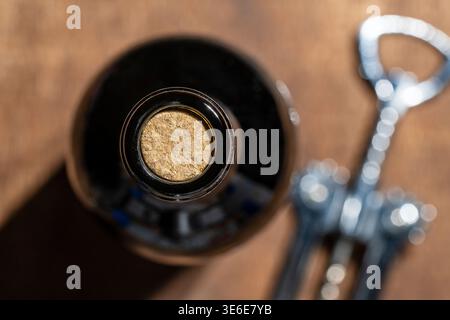 Nahaufnahme der Weinflasche aus dunklem Glas, die mit Naturkork verschlossen ist, neben einem Korkenzieher aus Metall auf einem hölzernen Hintergrund. Perfekt für Weinproben und Re Stockfoto