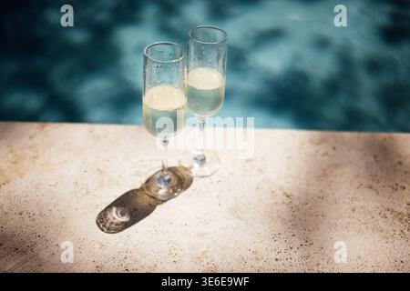 Zwei elegante Sektgläser mit prickelndem Getränk sitzen an einem Steinrand neben einem Swimmingpool und spiegeln das Wasser wider Stockfoto