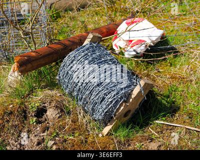 Rolle eines neuen, glänzenden Stacheldrahts für Zäune, der auf sonnendurchflutetem grünem Gras mit einem kaputten Holzpfosten und anderen weggeworfenen Materialien in einem ländlichen setti liegt Stockfoto