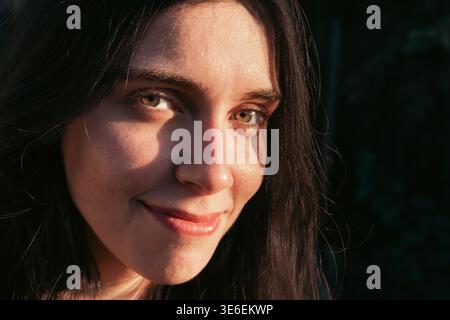 Nahaufnahme des Außenporträt einer jungen Frau mit einem sanften Lächeln in warmem natürlichem Sonnenlicht. Beauty- und Lifestyle-Bild mit natürlicher Hautstruktur, ausdrucksstark Stockfoto