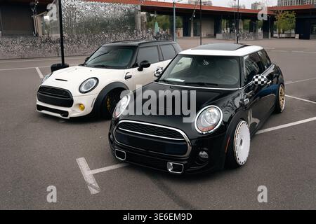 Blick auf zwei maßgeschneiderte Mini Cooper Autos, die nebeneinander auf einem offenen städtischen Parkplatz geparkt sind. Kompakte Fließhecken in kontrastierendem Schwarz und wh Stockfoto