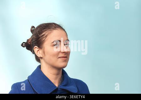 Junge Frau im blauen Mantel, die nachdenklich in einem sauberen Pastellporträt mit einem Haargutt aussieht Stockfoto