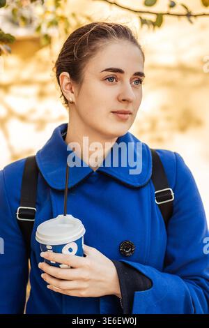 Eine junge Frau in einem blauen Mantel, die einen Kaffee zum Mitnehmen auf einer Herbststraße hält, ein nachdenkliches Porträt Stockfoto