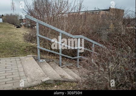 Nahaufnahme eines Sicherheitsgeländes auf einer Betontreppe im Freien, umgeben von trockenem Sträucher und entfernten Industriegebäuden für Sicherheit und Stadtverwaltung Stockfoto
