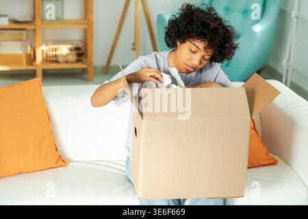 Afrikanisches Mädchen, das die Lieferung auspackt und in die Schachtel schaut. Glückliche Frau, die eine Schachtel öffnet. Eine Frau bekommt ein Paket, das sich die gelieferten Waren ansieht. Zufriedene Kunden positives Feedback, schnelle Expresslieferung Stockfoto