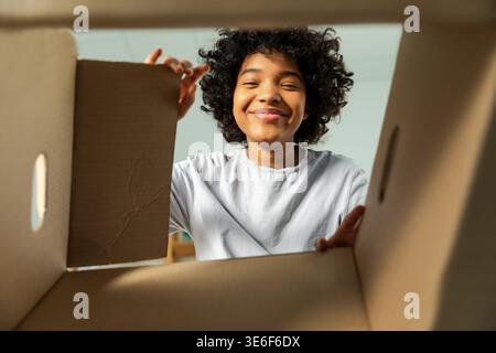 Ansicht von der Innenseite des Kartons. Afrikanisches Mädchen, das die Lieferung auspackt und in die Schachtel schaut. Frau öffnet Karton. Eine Frau bekommt ein Paket, das sich die gelieferten Waren ansieht. Zufriedenes positives Feedback des Kunden Stockfoto