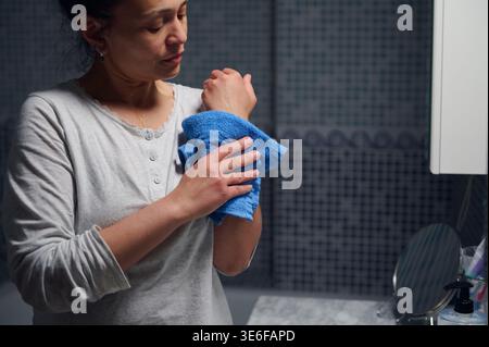 Eine Frau mittleren Alters im Pyjama wischt sich sanft mit einem blauen Handtuch am Waschbecken im Bad ab, während einer ruhigen morgendlichen Hygiene- und Schönheitsroutine betont Stockfoto